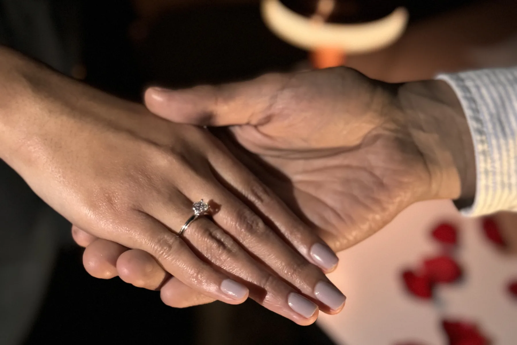 Manos de una pareja con anillo de compromiso, a la luz de una vela y entre pétalos de rosa